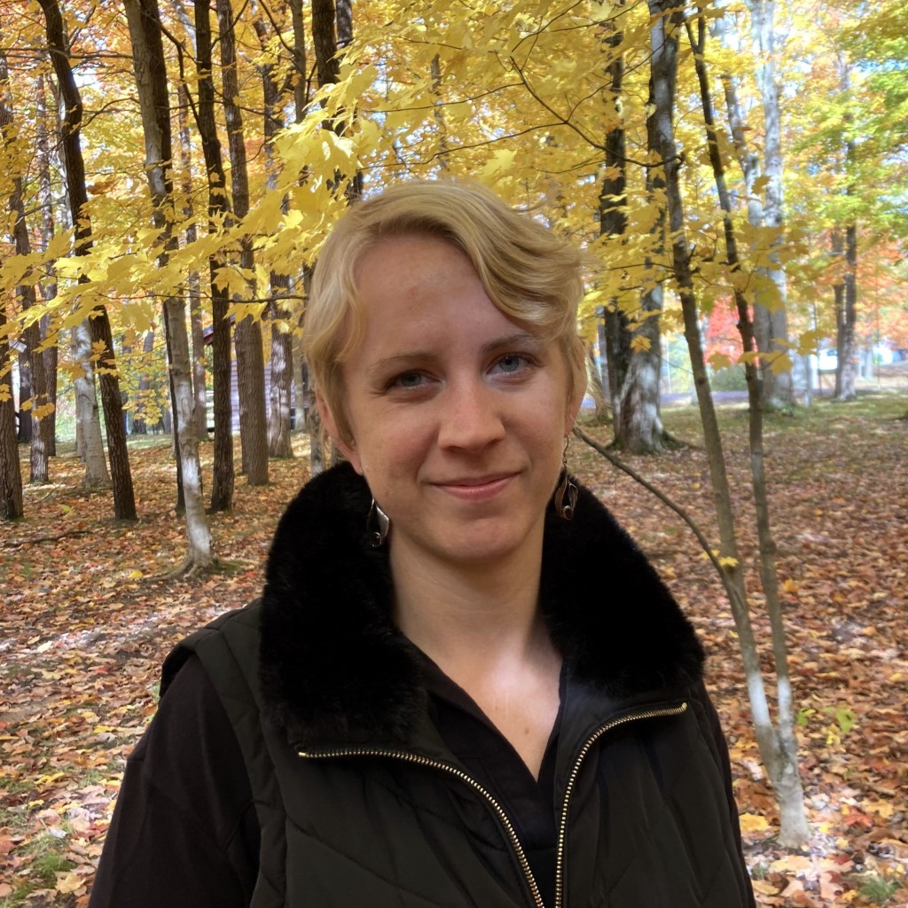 Woman with short blonde hair with a knowing expression standing in a fall forest